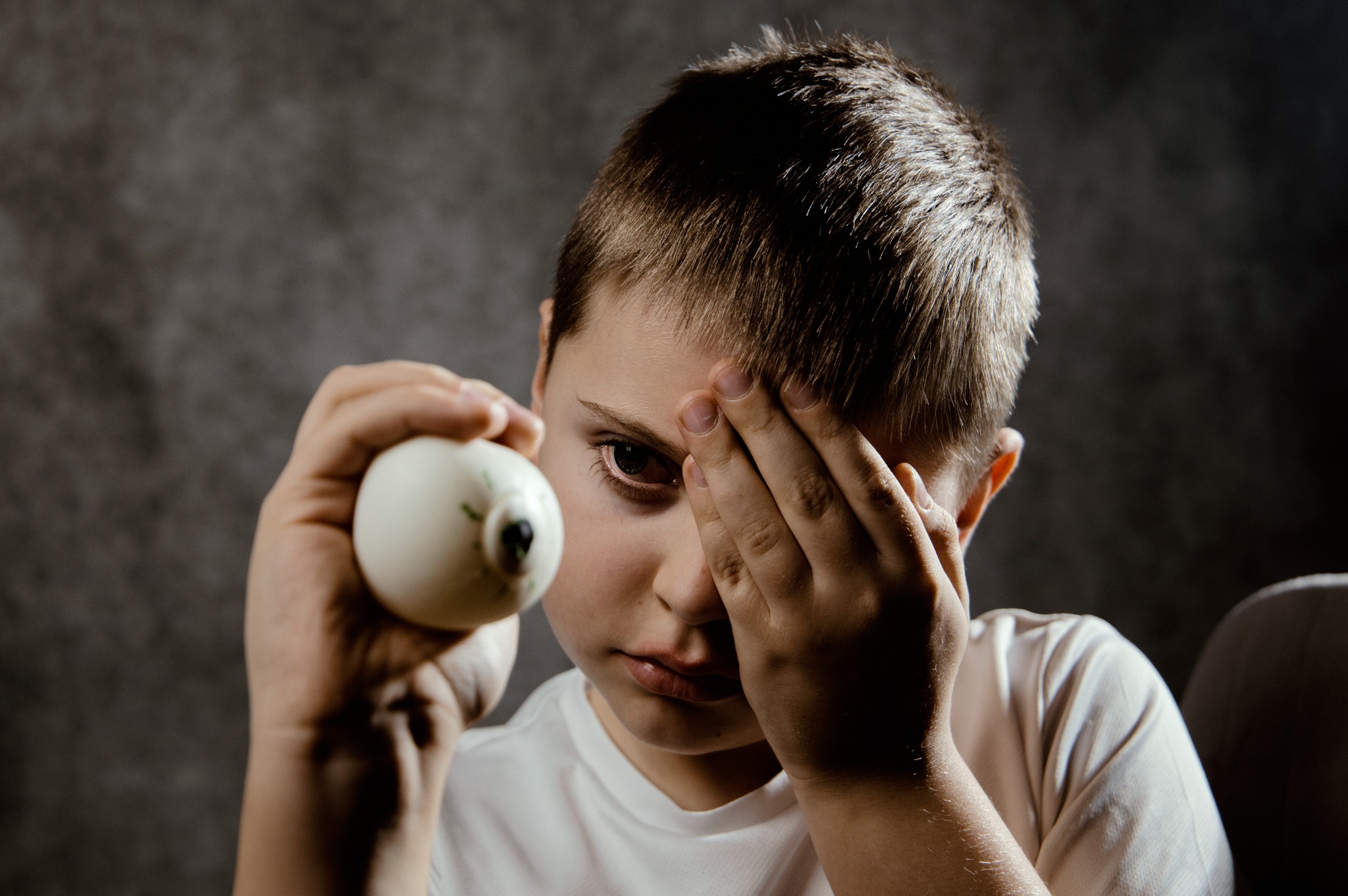 Portret chłopca z dekoracyjnym jajkiem w dłoni, zasłaniającego oko drugą ręką. Ciemne tło, oświetlenie skupione na twarzy. Ujęcie z bliska, widoczny wyraz zamyślenia.