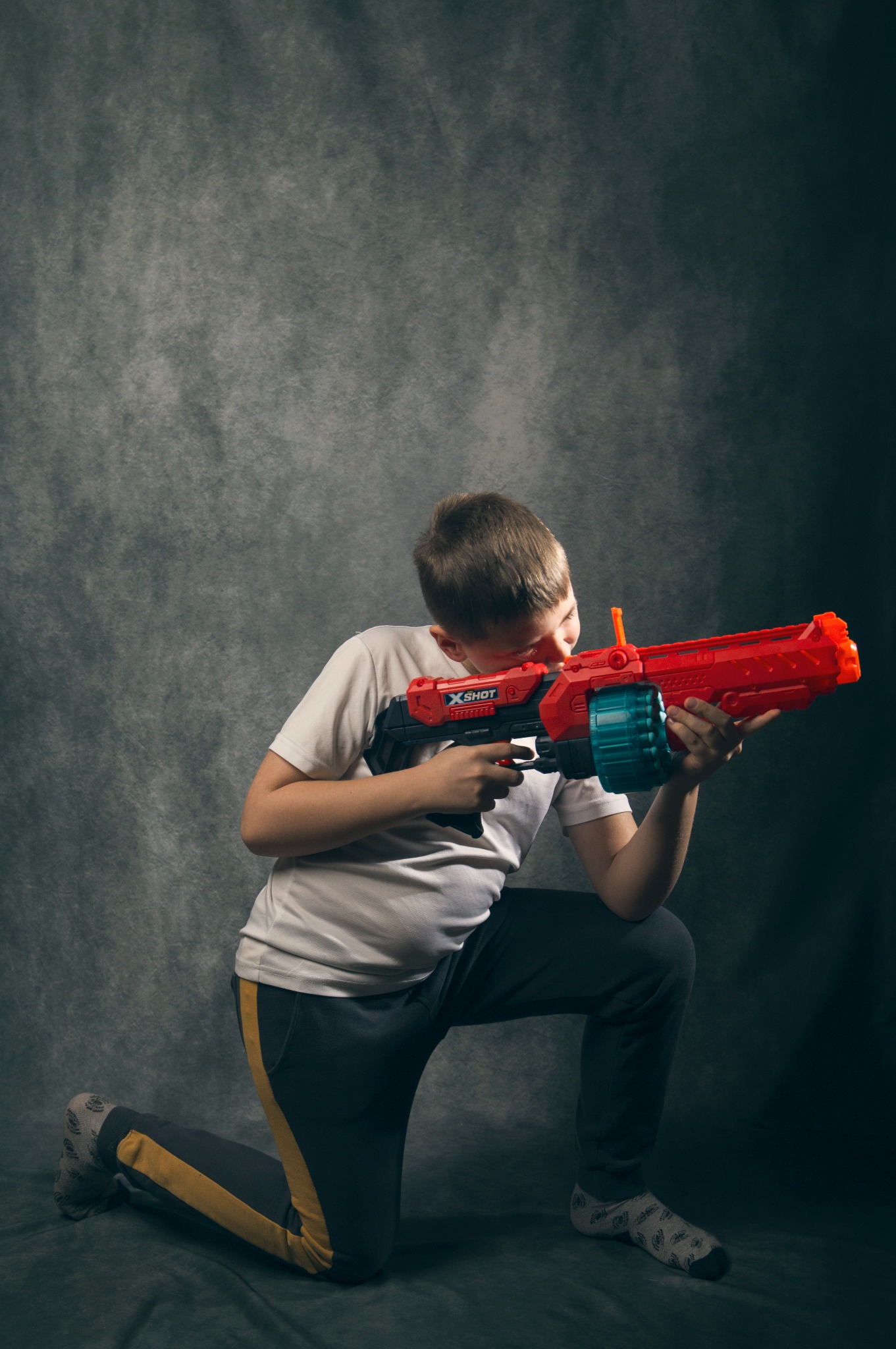 Chłopiec klęczy, celując z czerwono-czarnego pistoletu X-Shot z turkusowym magazynkiem na tle szarego tła. Ma na sobie białą koszulkę i ciemne spodnie z żółtym paskiem.
