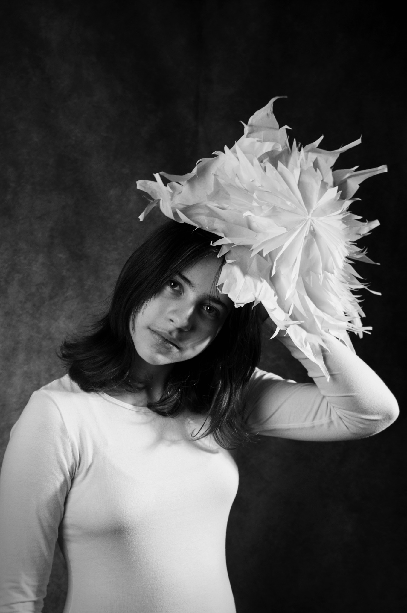 Czarno-biały portret młodej kobiety z dużym, papierowym kwiatem na głowie, spoglądającej w dół, na ciemnym tle.