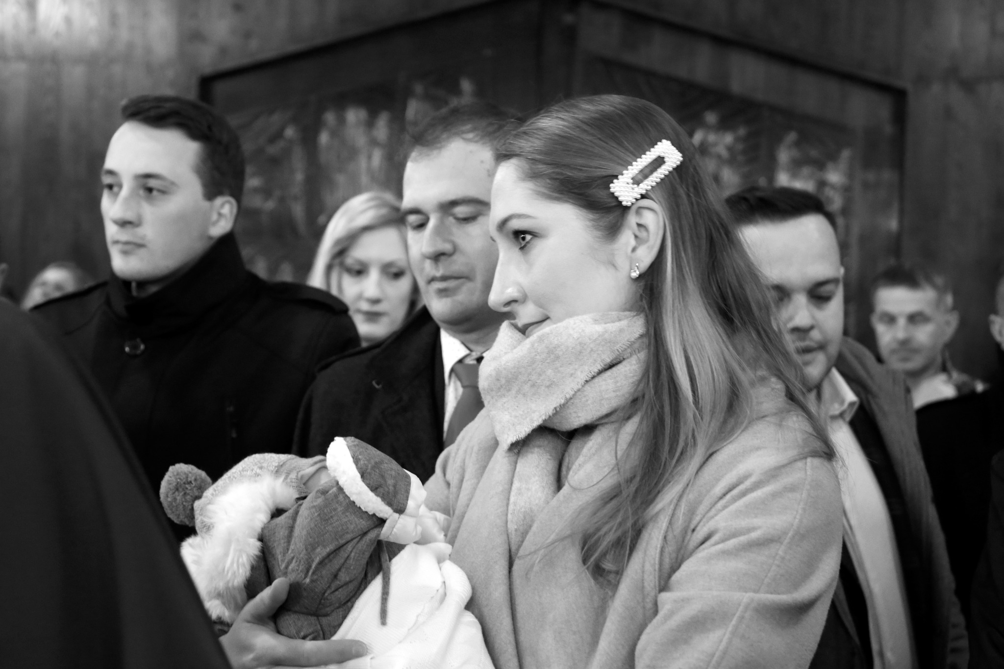 Czarno-białe ujęcie z uroczystości chrztu: kobieta z ozdobną spinką we włosach trzyma dziecko, w tle widoczni goście i fragment obrazu na ścianie kościoła.