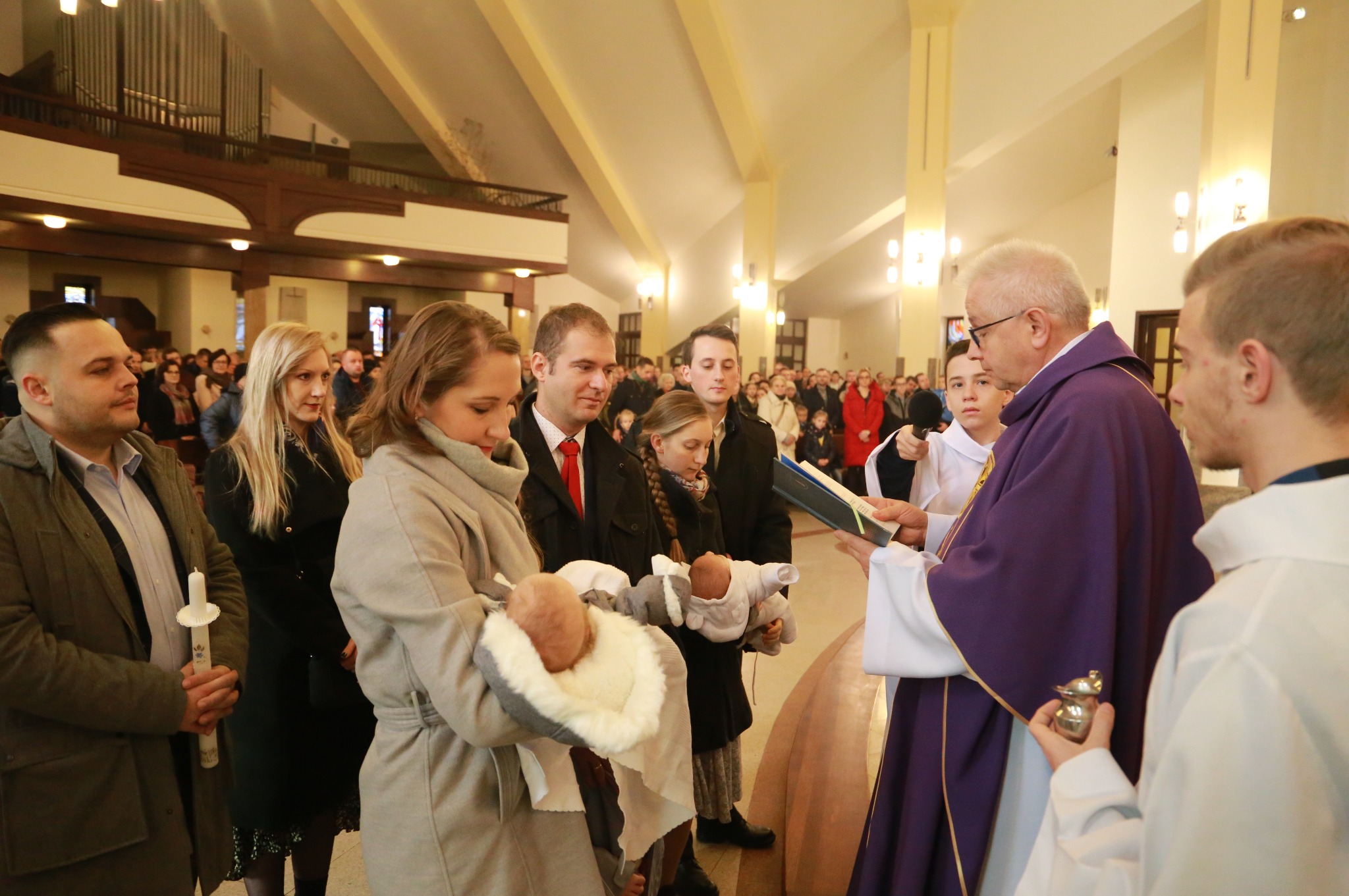 Uroczystość chrztu w kościele: ksiądz w fioletowej szacie błogosławi niemowlęta trzymane przez rodziców, w tle zgromadzeni goście i architektoniczne detale wnętrza.