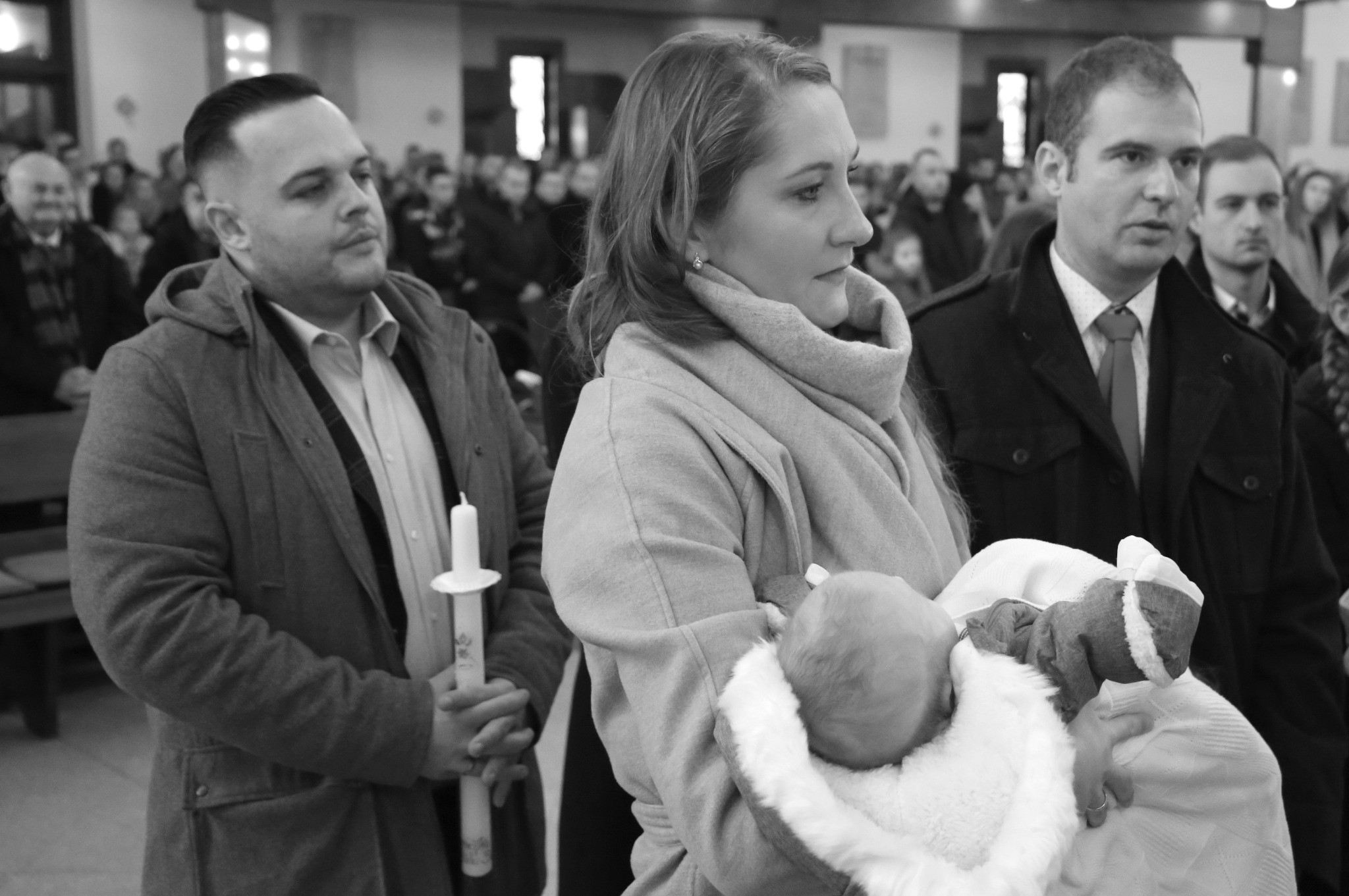 Czarno-białe zdjęcie z ceremonii chrztu: kobieta trzyma niemowlę, obok mężczyzna z świecą, w tle zgromadzeni ludzie w kościele.