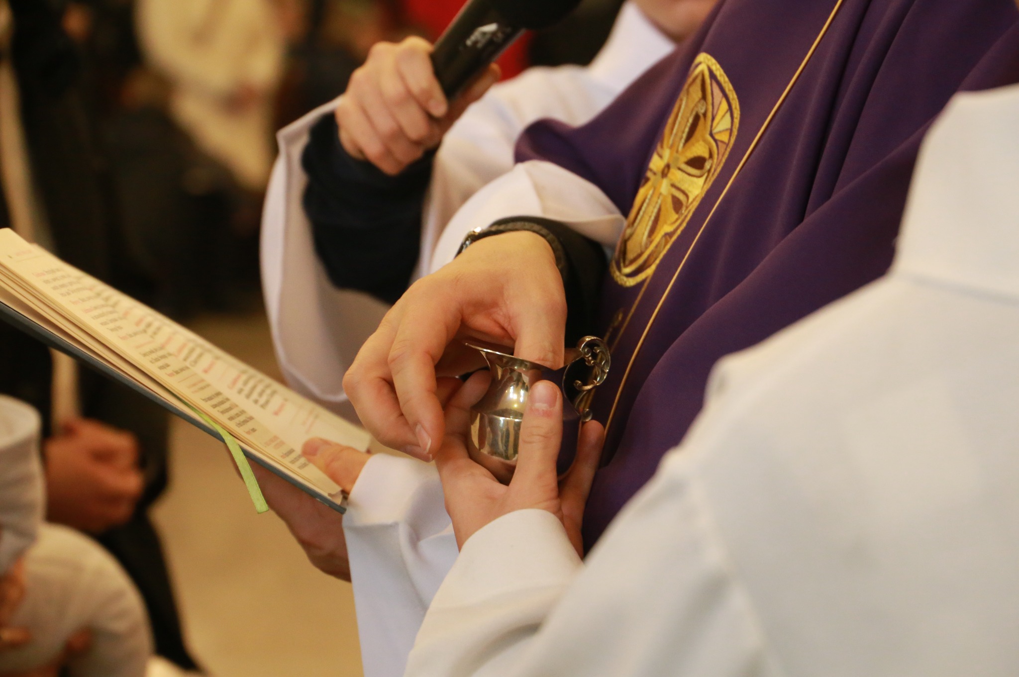 Detal z ceremonii religijnej: ksiądz w fioletowej szacie z haftowanym symbolem, trzymający srebrny dzbanuszek. W tle otwarta księga i mikrofon.