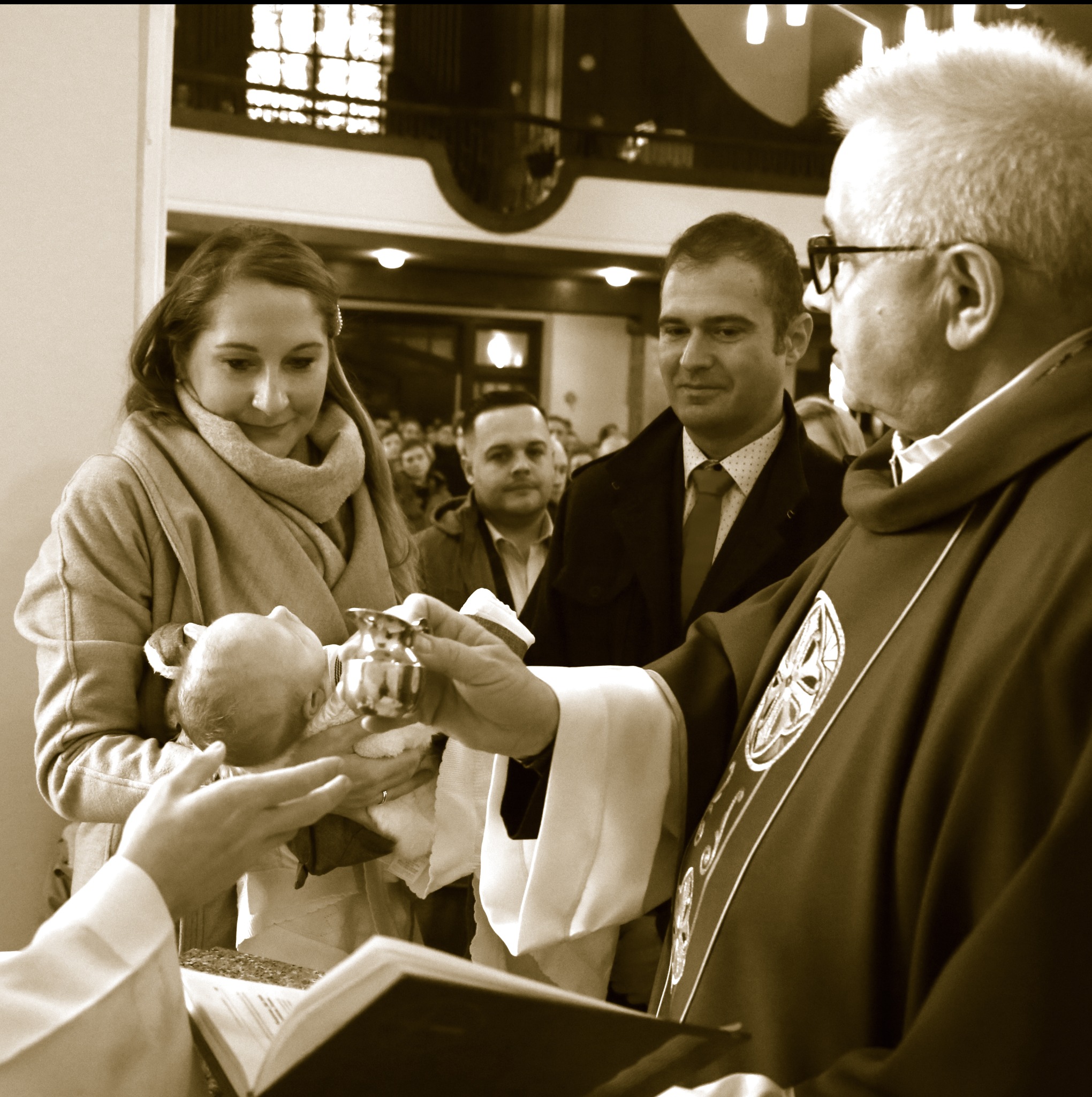 Sepiowe ujęcie z ceremonii chrztu: matka trzyma dziecko, ksiądz polewa wodą, w tle rodzina w kościele. Zbliżenie na sakrament z elementami architektury sakralnej.