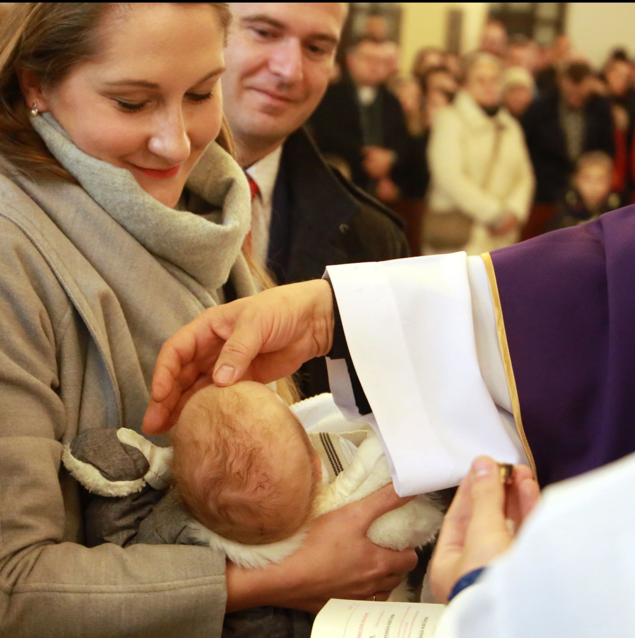 Chrzest dziecka: matka trzyma niemowlę, ksiądz błogosławi. W tle widoczni goście w kościele, delikatne światło podkreśla uroczystość.