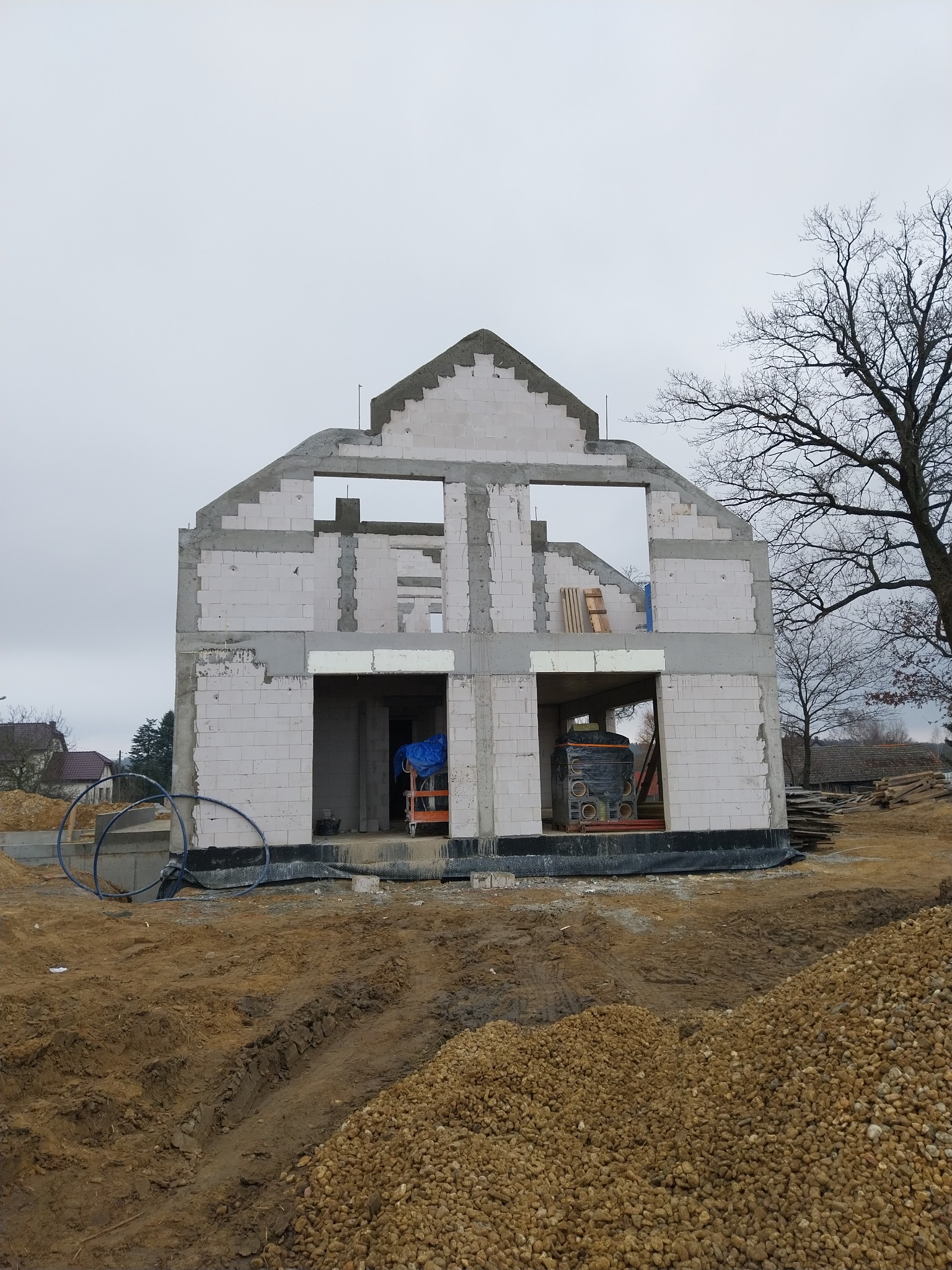 Surowy stan domu z białych bloczków, widoczne otwory okienne i drzwiowe, na tle ziemi i drzew. Etap budowy z elementami betonowymi i materiałami budowlanymi wewnątrz.
