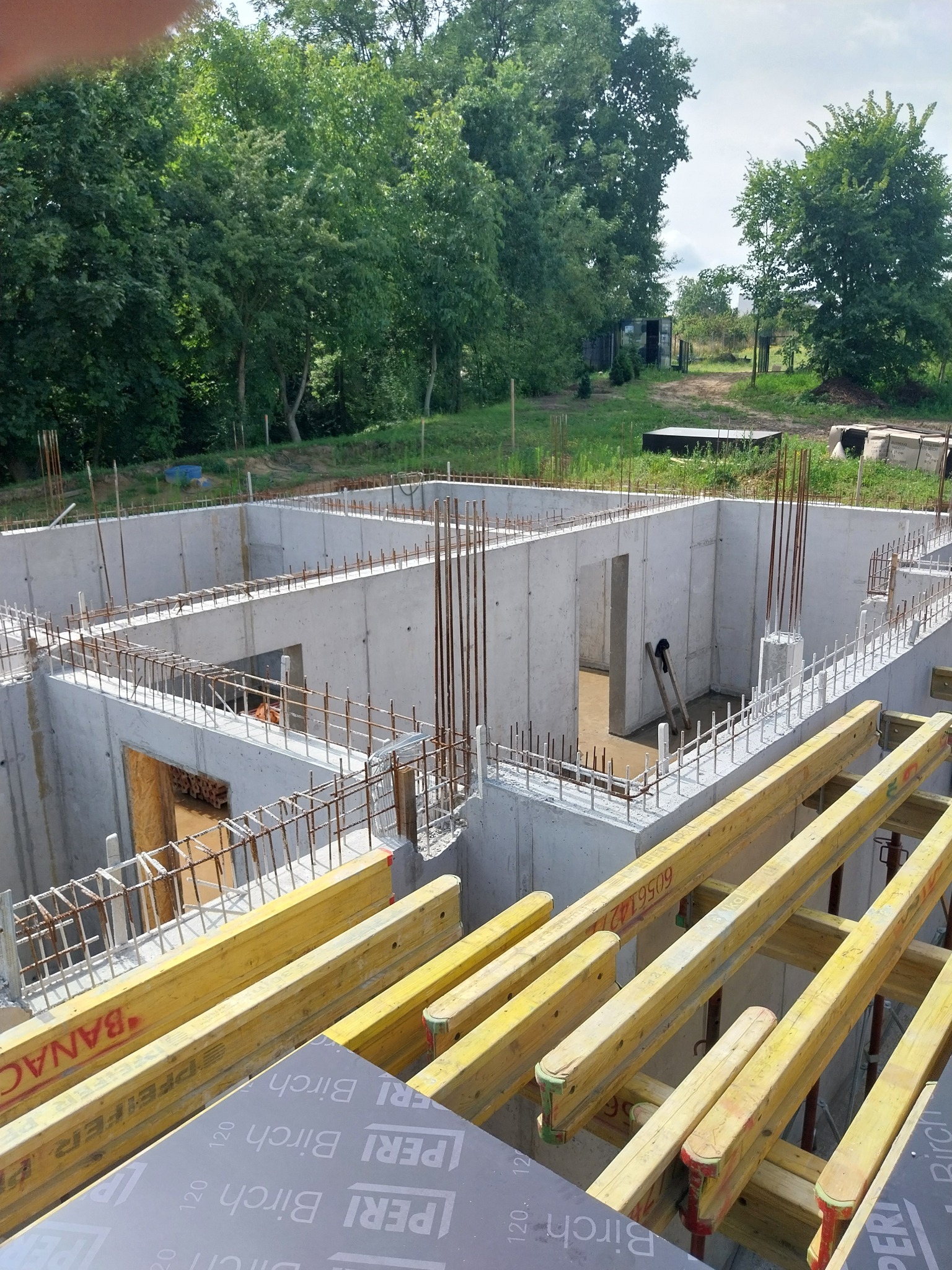 Żelbetowe ściany fundamentowe w trakcie budowy, zbrojenie stalowe, szalunki PERI BIRCH, widok z góry na plac budowy otoczony zielenią.