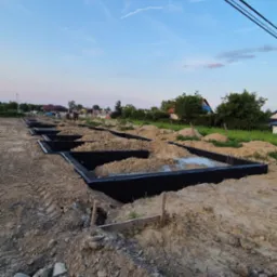 Fundamenty domu jednorodzinnego, zabezpieczone czarną folią hydroizolacyjną, widoczne na placu budowy w słoneczny dzień. W tle zabudowa jednorodzinna i niebo.