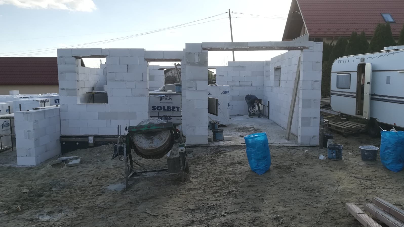 Budowa domu z białych bloczków Solbet, widoczna betoniarka i pracownik przy zbrojeniu wewnątrz surowej konstrukcji. W tle przyczepa kempingowa i dom sąsiada.