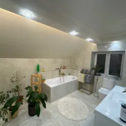 Jasna łazienka na poddaszu z wanną, umywalką i toaletą. Beżowe płytki imitujące marmur, rośliny doniczkowe, złote akcenty i oświetlenie punktowe.