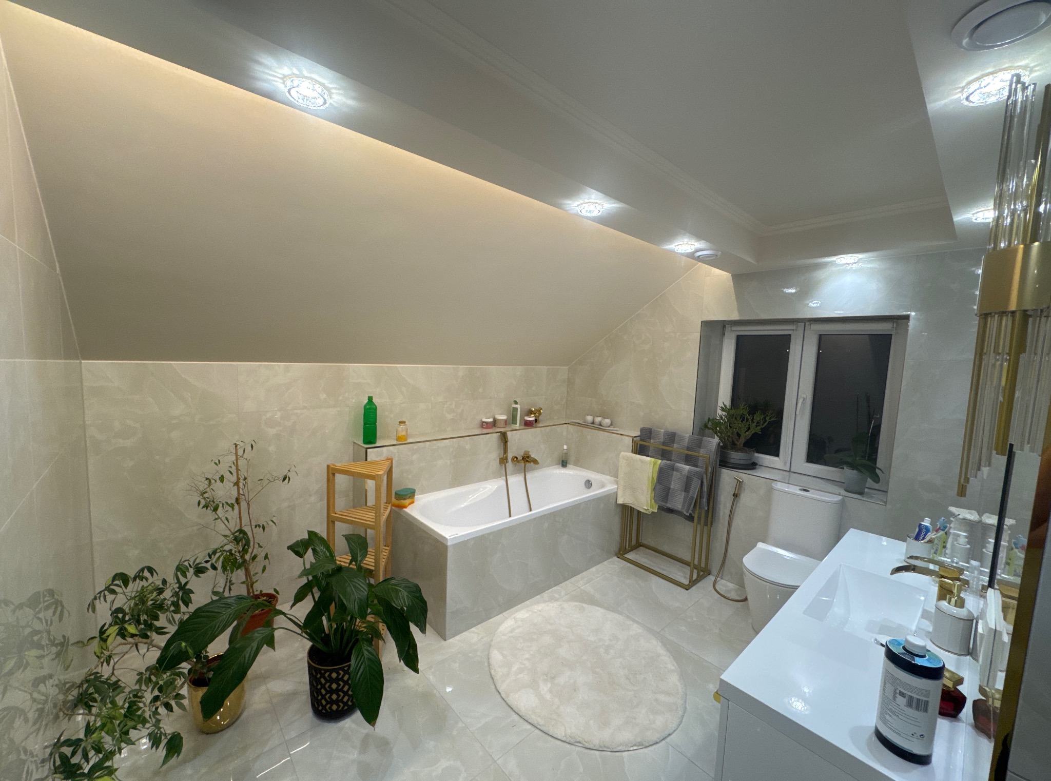 Jasna łazienka na poddaszu z wanną, umywalką i toaletą. Beżowe płytki imitujące marmur, rośliny doniczkowe, złote akcenty i oświetlenie punktowe.
