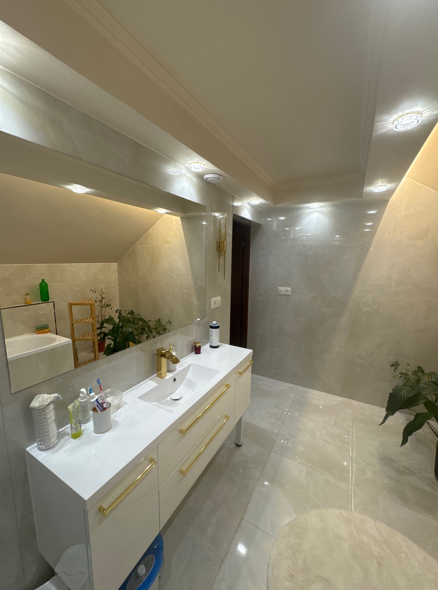 Jasna łazienka z umywalką na szafce z złotymi uchwytami, lustrem i roślinami. Marmurowe płytki na ścianach i podłodze, punktowe oświetlenie sufitowe.