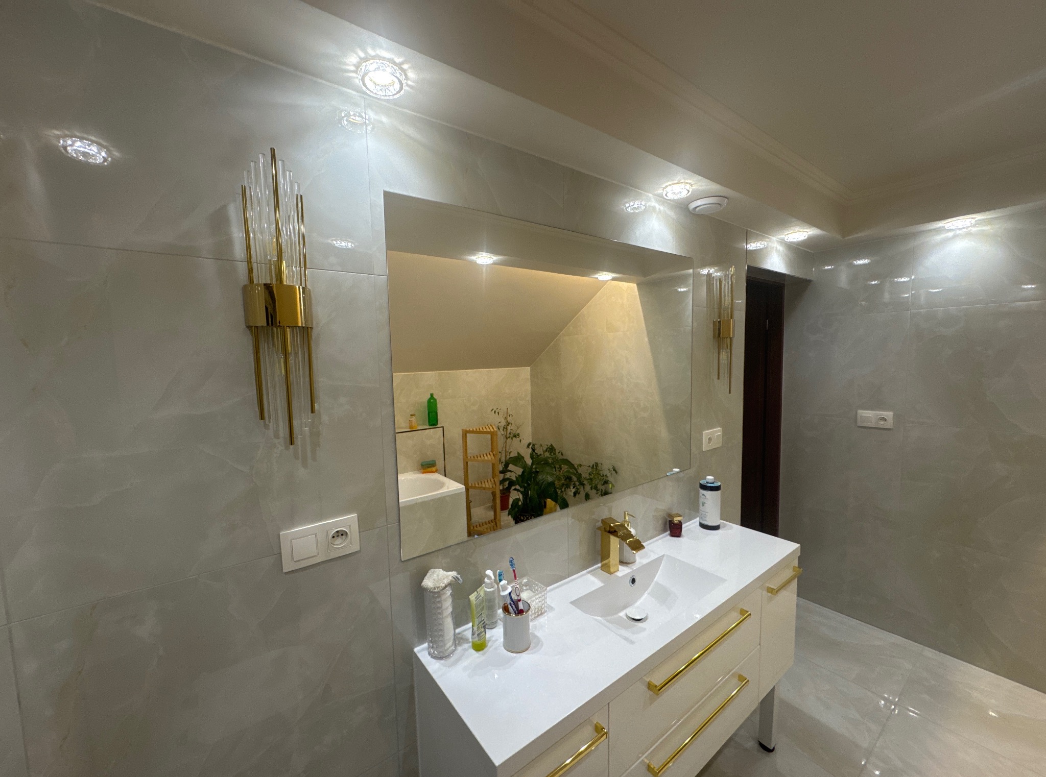 Jasna łazienka z lustrem odbijającym fragment wanny i rośliny. Biała szafka pod umywalką z złotymi uchwytami. Na ścianie kinkiet w stylu art deco.