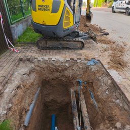 OPTiTEK USŁUGI WOD-KAN Ełk - Wykop z widocznymi rurami wodociągowymi i kanalizacyjnymi, częściowo odsłoniętymi, z żółtą mini koparką Wacker Neuson w tle i zabezpieczeniami wykopu.