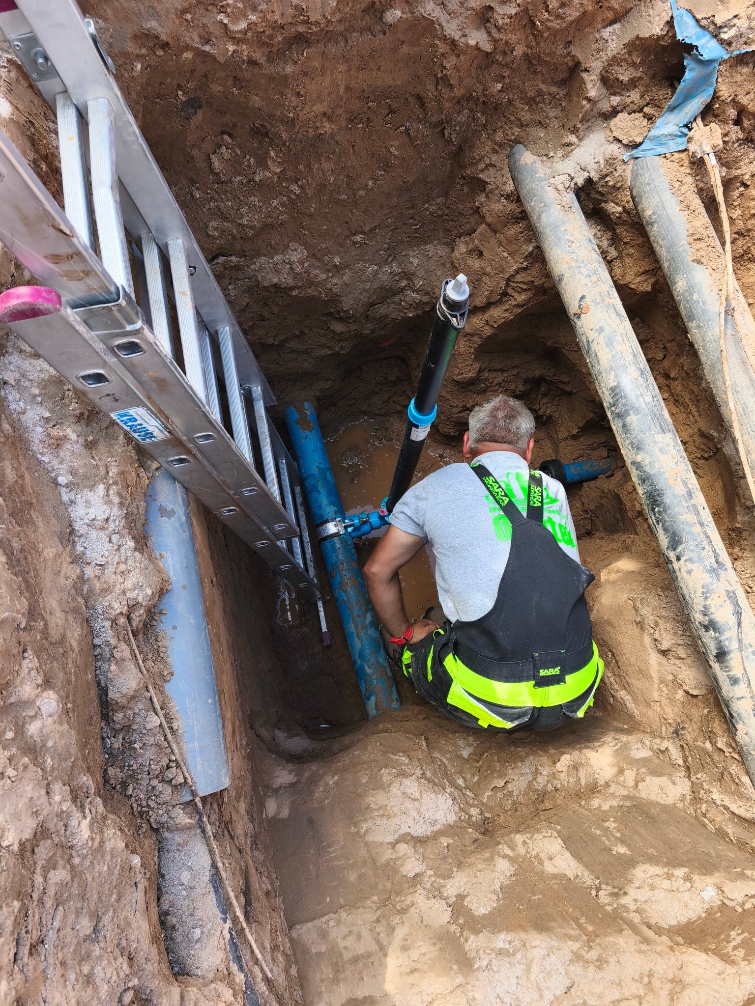 Pracownik w wykopie łączy niebieskie rury przy pomocy złączek. Widoczna drabina, inne rury i ziemia. Prace instalacyjne przyłącza wod-kan.