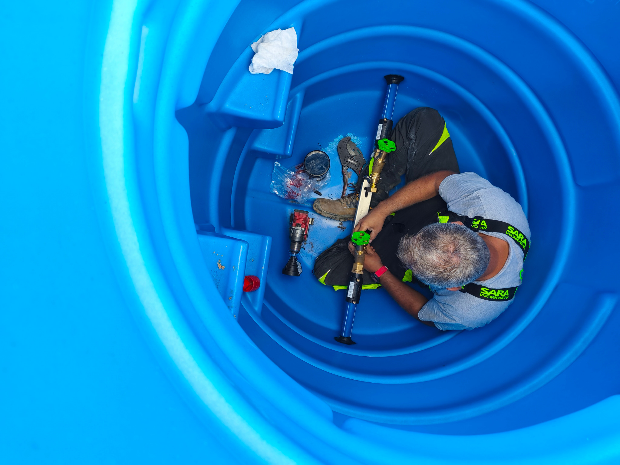Instalator wewnątrz niebieskiego zbiornika, montuje rurę z zaworem. Widoczne narzędzia, buty robocze i akcesoria. Ujęcie z góry.
