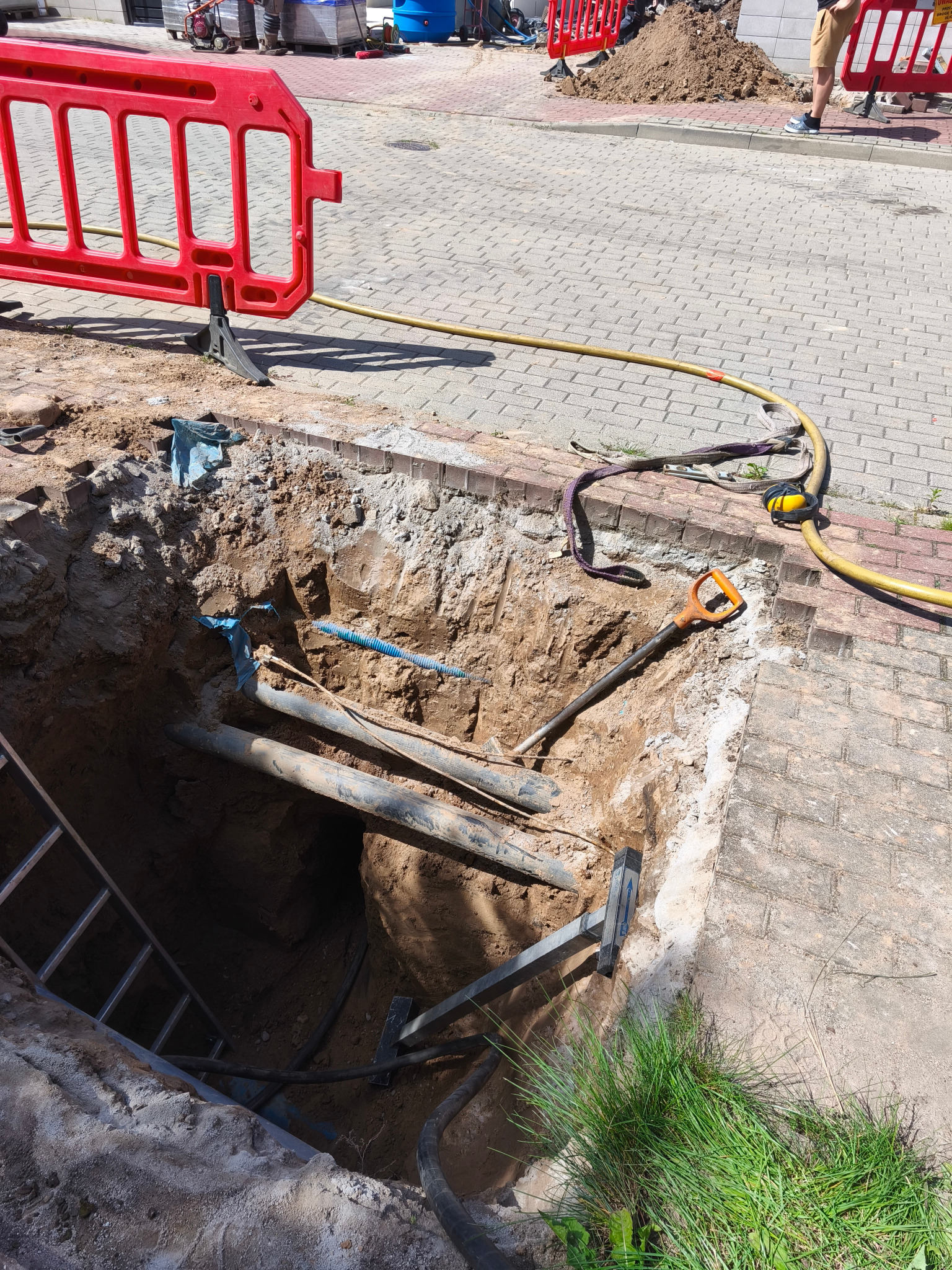 Odkopane rury w wykopie na chodniku, zabezpieczone barierkami i drabiną, narzędzia pracy obok. Prace ziemne przyłącza wod-kan w trakcie realizacji.