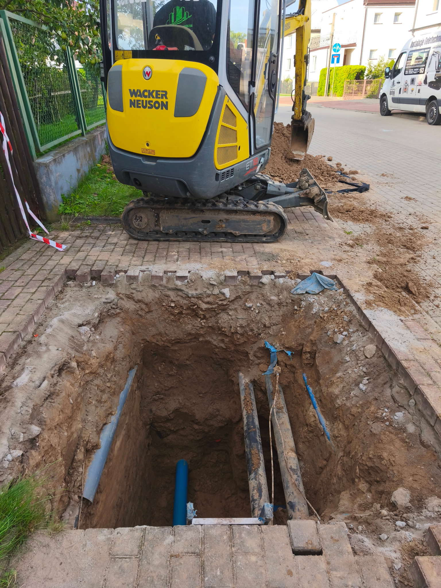 Wykop z widocznymi rurami wodociągowymi i kanalizacyjnymi, częściowo odsłoniętymi, z żółtą mini koparką Wacker Neuson w tle i zabezpieczeniami wykopu.