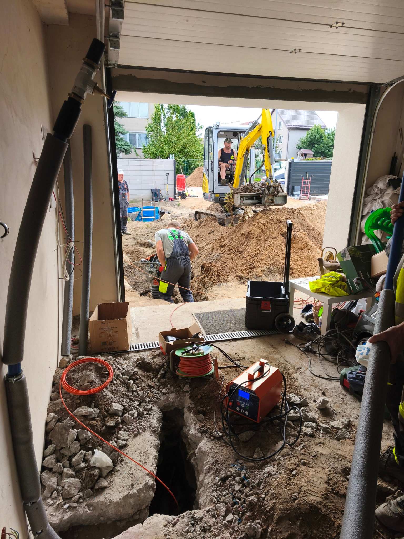 Prace ziemne z użyciem minikoparki i zagęszczarki w garażu, widoczne rury w izolacji, sprzęt spawalniczy i wykop pod przyłącze. Prace instalacyjne w toku.