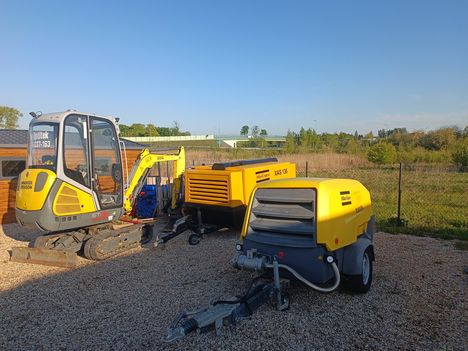 Żółty minikoparka Wacker Neuson ET18 i dwa kompresory Atlas Copco XAS 136 na żwirowym podłożu, w tle zieleń i błękitne niebo.