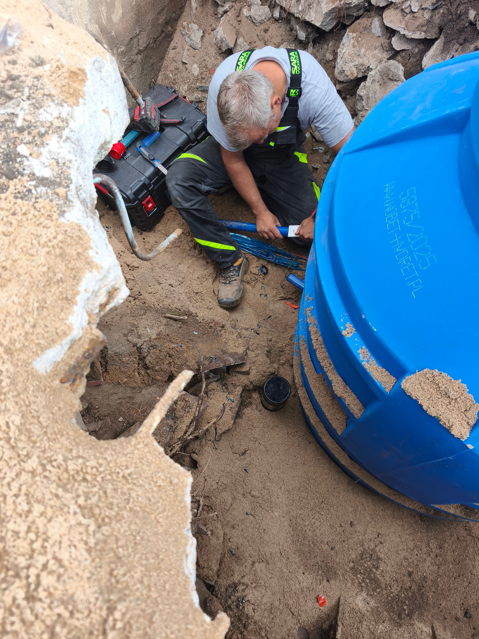 Monter w wykopie łączy rury do niebieskiego zbiornika. Widoczna skrzynka narzędziowa, ziemia i fragment betonowej konstrukcji. Prace instalacyjne w toku.
