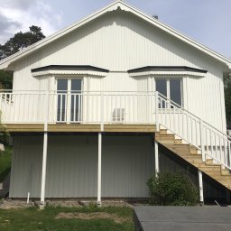 SMART4LIFE PROSTA SPÓŁKA AKCYJNA - Biały dom szkieletowy z drewnianymi schodami i balkonem, widok z dołu, elementy dekoracyjne okien, jasne oświetlenie, zielone otoczenie.