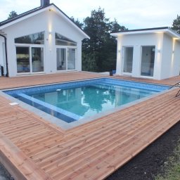 SMART4LIFE PROSTA SPÓŁKA AKCYJNA - Nowoczesny dom z basenem otoczonym tarasem z desek. Widoczne białe elewacje budynków i błękitna woda w basenie. Dwa leżaki stoją obok basenu.