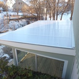 SMART4LIFE PROSTA SPÓŁKA AKCYJNA - Zadaszony taras z przezroczystym poliwęglanem, widok z góry. Drewniana podłoga tarasu widoczna przez szklane drzwi. W tle ośnieżony ogród i domy.