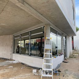 SMART4LIFE PROSTA SPÓŁKA AKCYJNA - Montaż okien w surowym budynku z betonu komórkowego. Widoczna drabina, narzędzia i częściowo zamontowane okna z czarnymi ramami. W tle zaparkowany biały bus.