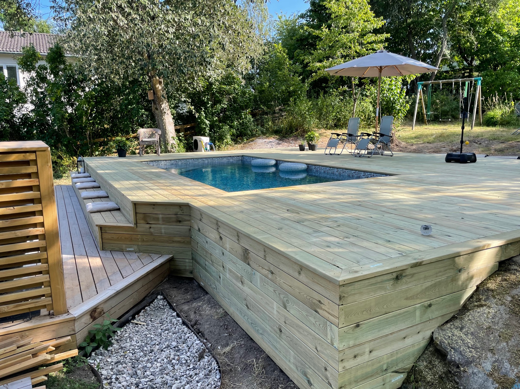 Nowoczesny taras z drewna otaczający basen, z leżakami i parasolem, w tle zieleń ogrodu. Widoczne schody i oświetlenie tarasowe.
