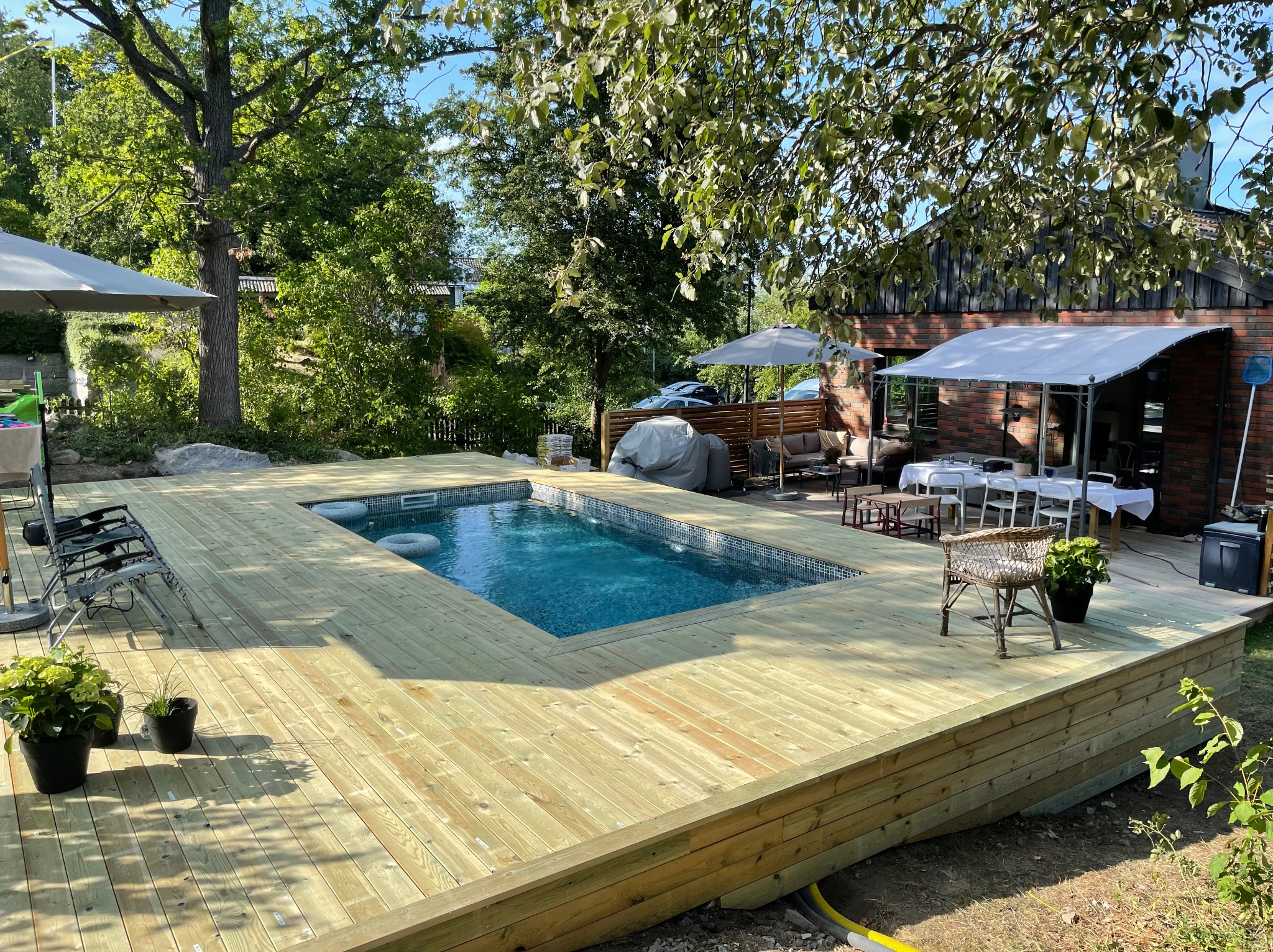 Jasny taras drewniany otaczający basen z mozaiką, z krzesłem wiklinowym i roślinami w donicach. W tle ogród i budynek z cegły.