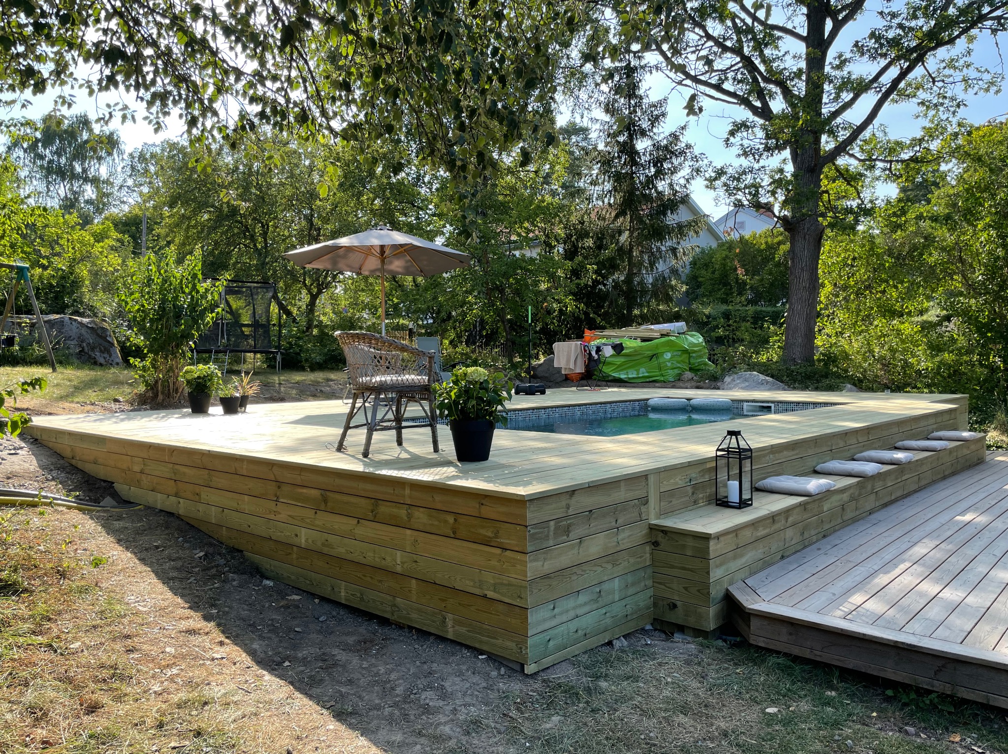 Nowoczesny, drewniany taras z basenem, otoczony zielenią. W tle parasol i krzesło z wikliny. Całość w jasnych, naturalnych barwach, z delikatnym oświetleniem.
