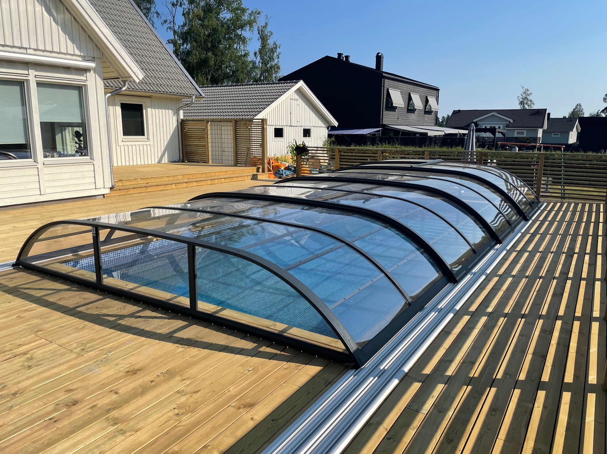 Zadaszenie basenu z poliwęglanu na drewnianym tarasie, widoczne domy w tle. Słoneczny dzień, błękitna woda w basenie. Nowoczesna architektura.
