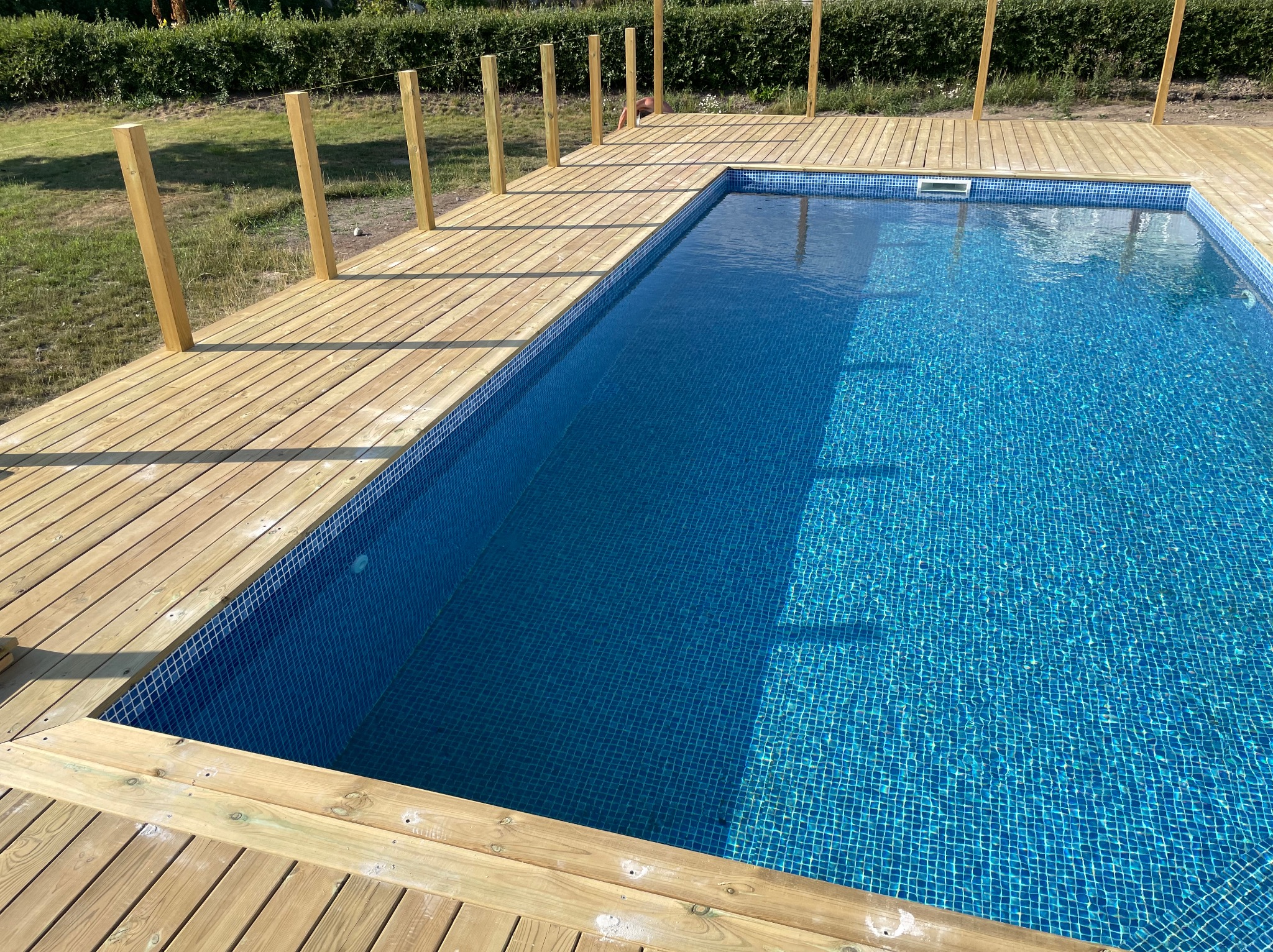 Narożny basen z błękitną mozaiką, otoczony jasną drewnianą konstrukcją. Widoczne słupki zabezpieczające. Trawa i roślinność w tle. Słoneczny dzień.