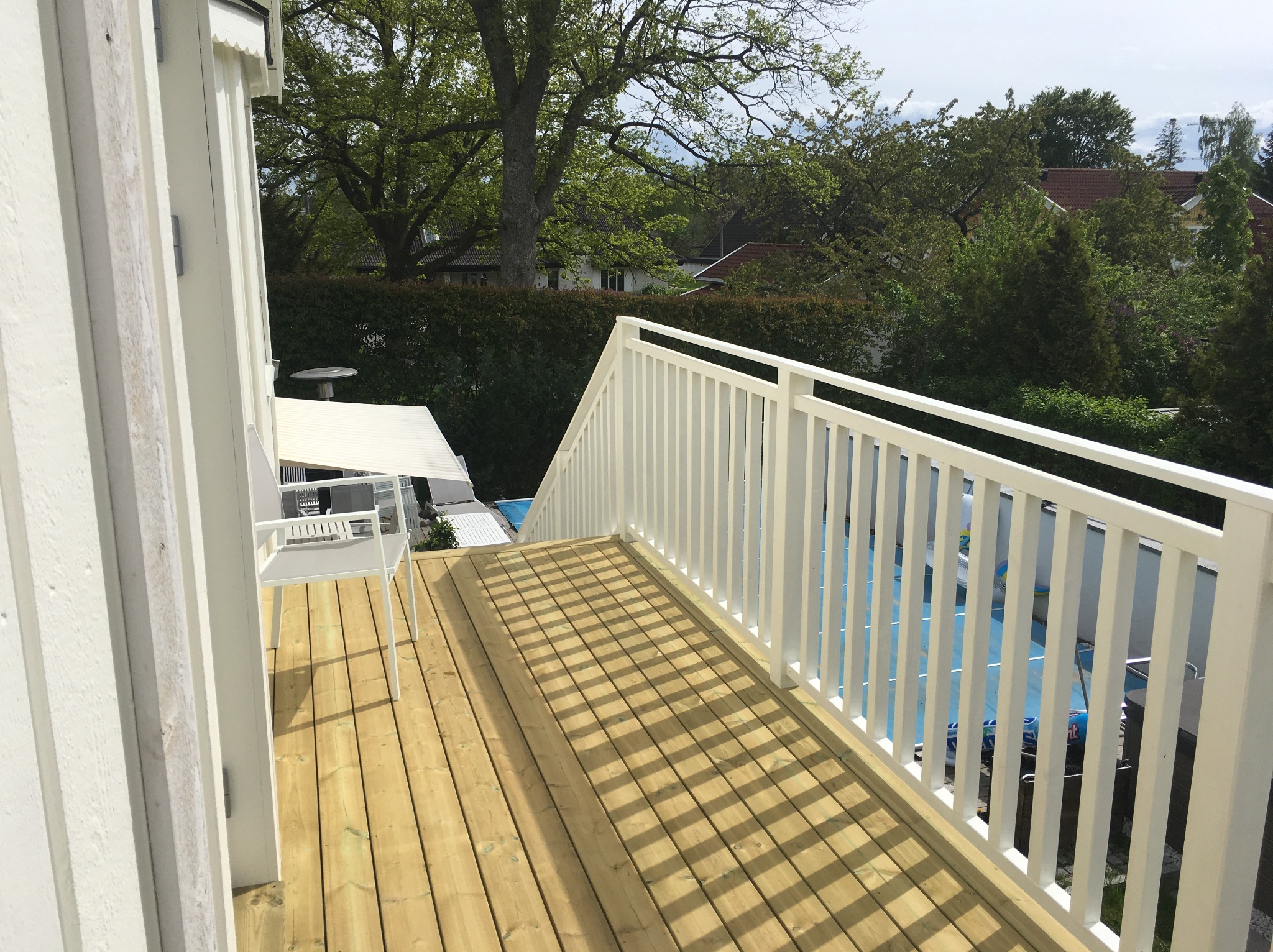 Nowy drewniany taras z białą balustradą, widok z góry, z meblami ogrodowymi w tle i fragmentem basenu. Cień tworzy wzór na deskach. Jasne, naturalne oświetlenie.