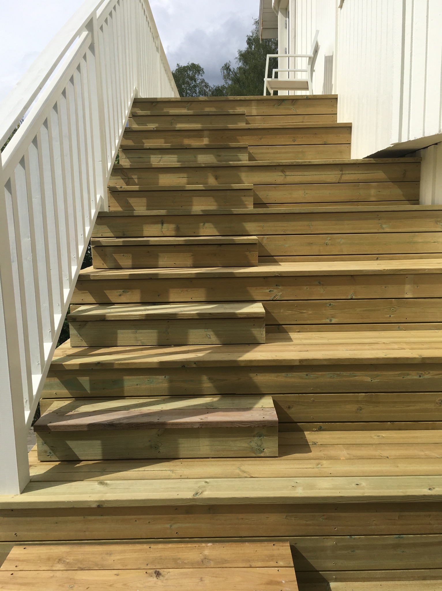Drewniane schody zewnętrzne z białymi balustradami prowadzące do budynku. Widoczne poziome deski i cień. Jasne, naturalne drewno.
