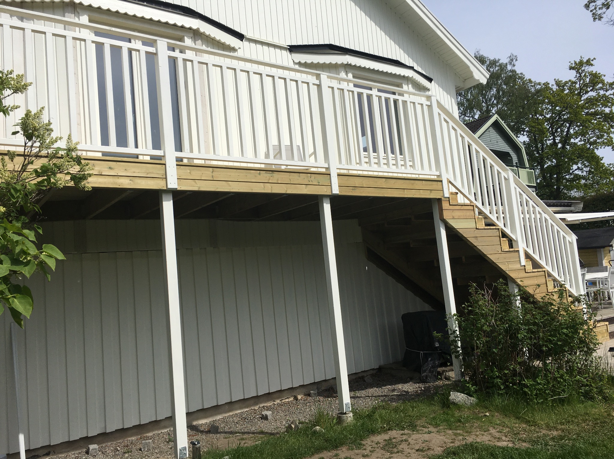 Drewniany taras z białą balustradą przy domu z jasną elewacją. Widoczne schody prowadzące na taras i podpory konstrukcji. Tło stanowi zieleń drzew i niebieskie niebo.