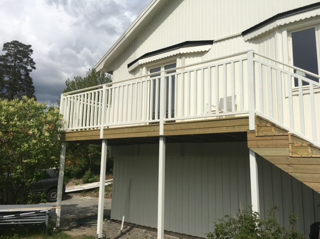 Biały, drewniany taras z balustradą i schodami przy domu z jasną elewacją. Widok z dołu, fragment ogrodu z zielenią. Pochmurne niebo w tle.