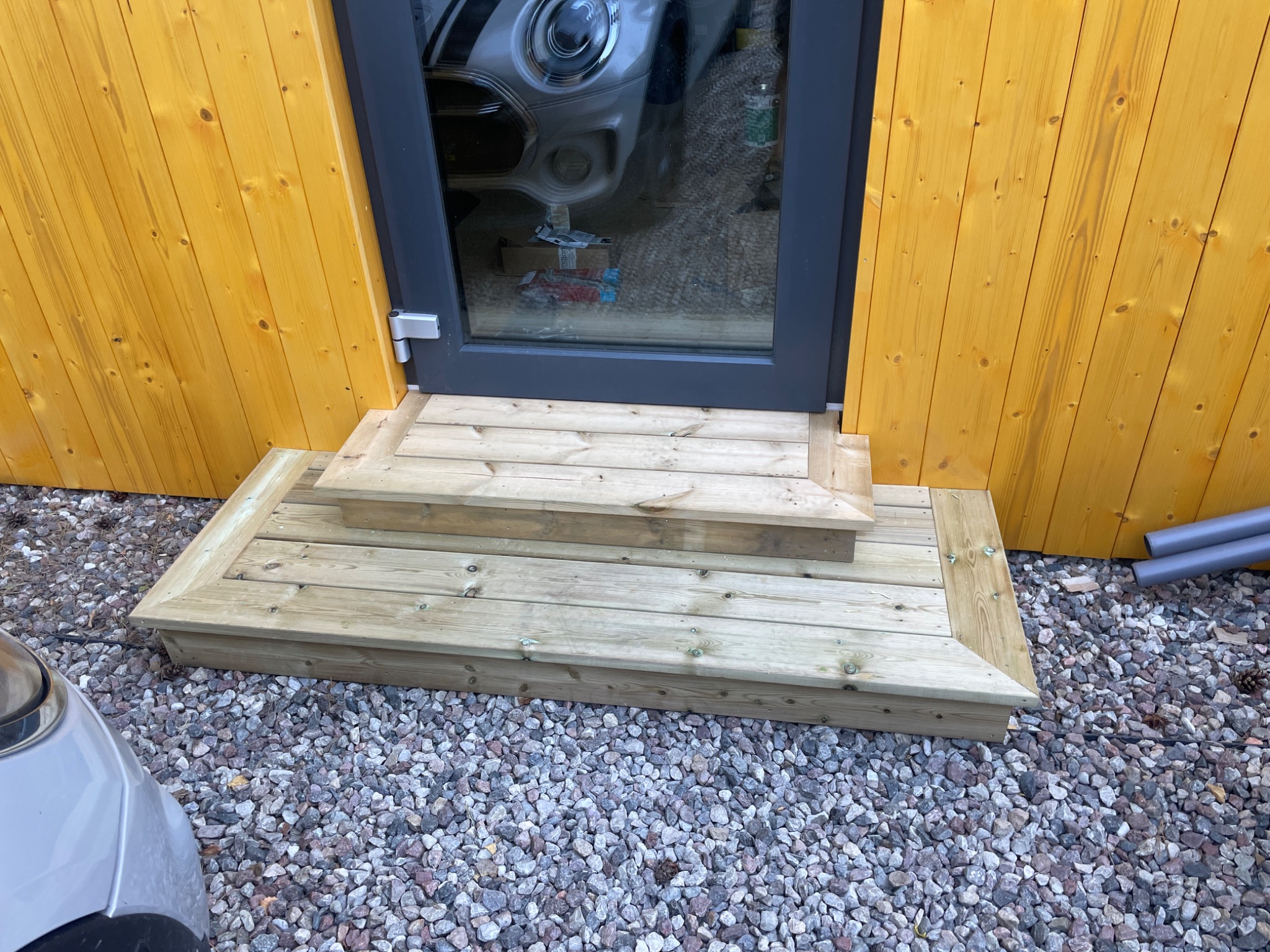 Drewniane schody wejściowe prowadzące do drzwi z szarą ramą, widoczne na tle żółtej elewacji. Podłoże wyłożone drobnymi kamieniami.