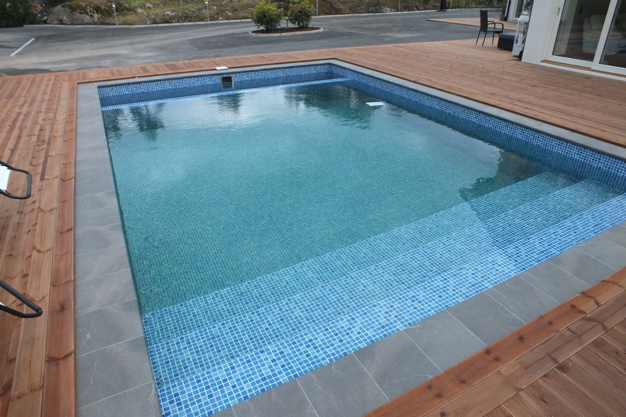 Narożne ujęcie turkusowego basenu z mozaiką, otoczonego drewnianym tarasem, z szarą obwódką i schodkami wewnątrz. W tle zieleń i fragment parkingu.