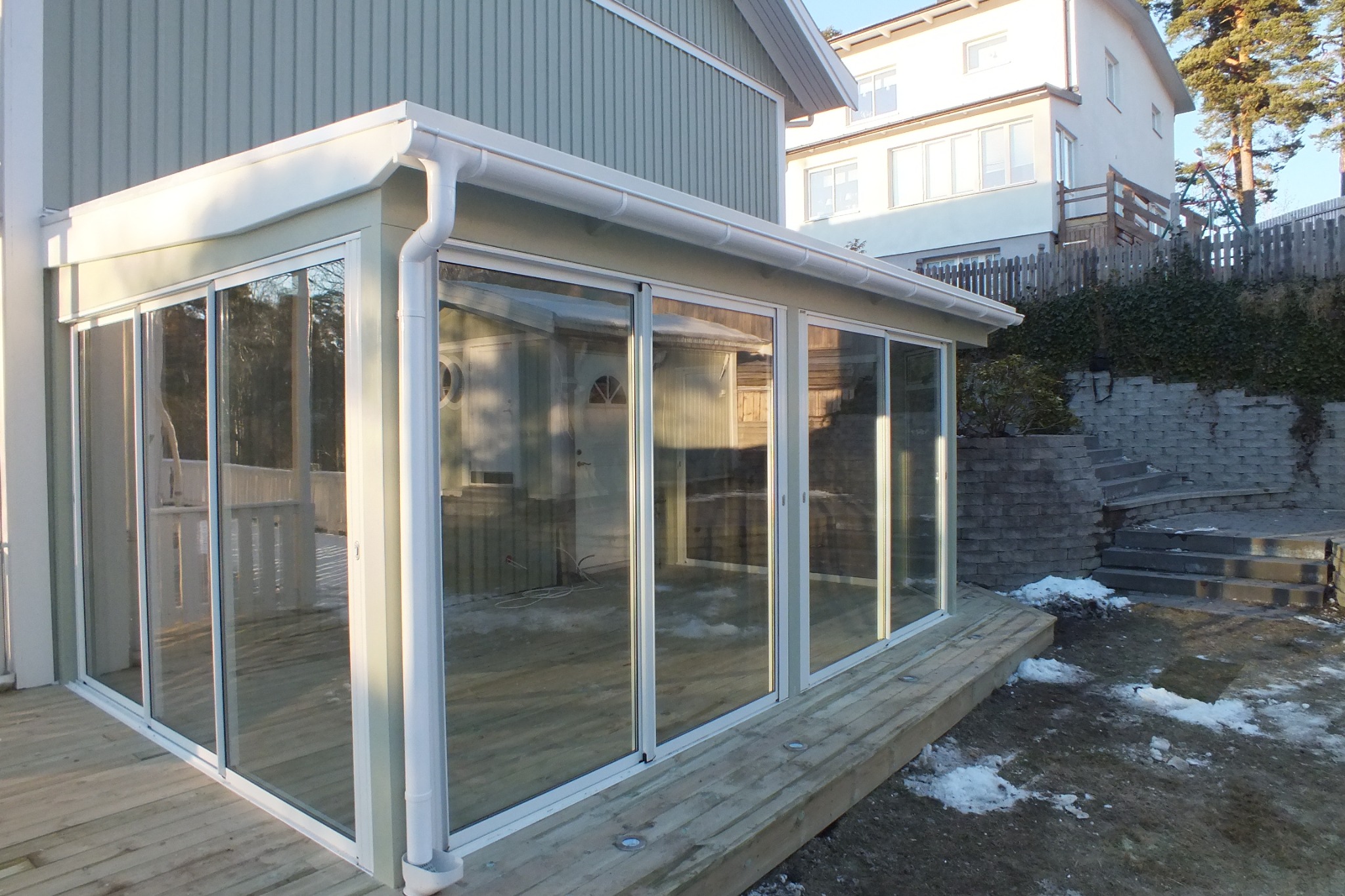 Zadaszony taras z przesuwnymi oknami aluminiowymi i drewnianą podłogą, przylegający do domu z jasną elewacją. Widoczne elementy ogrodu i schody kamienne.