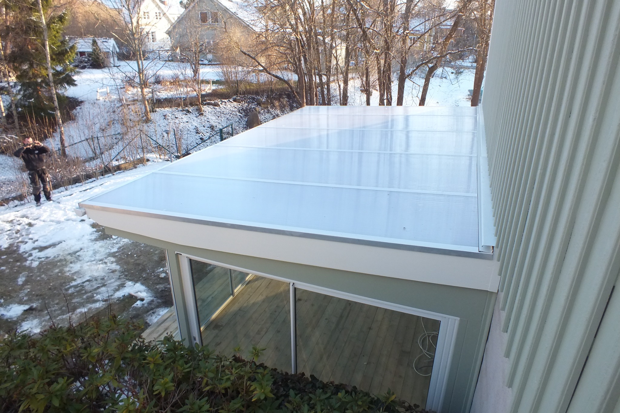 Zadaszony taras z przezroczystym poliwęglanem, widok z góry. Drewniana podłoga tarasu widoczna przez szklane drzwi. W tle ośnieżony ogród i domy.