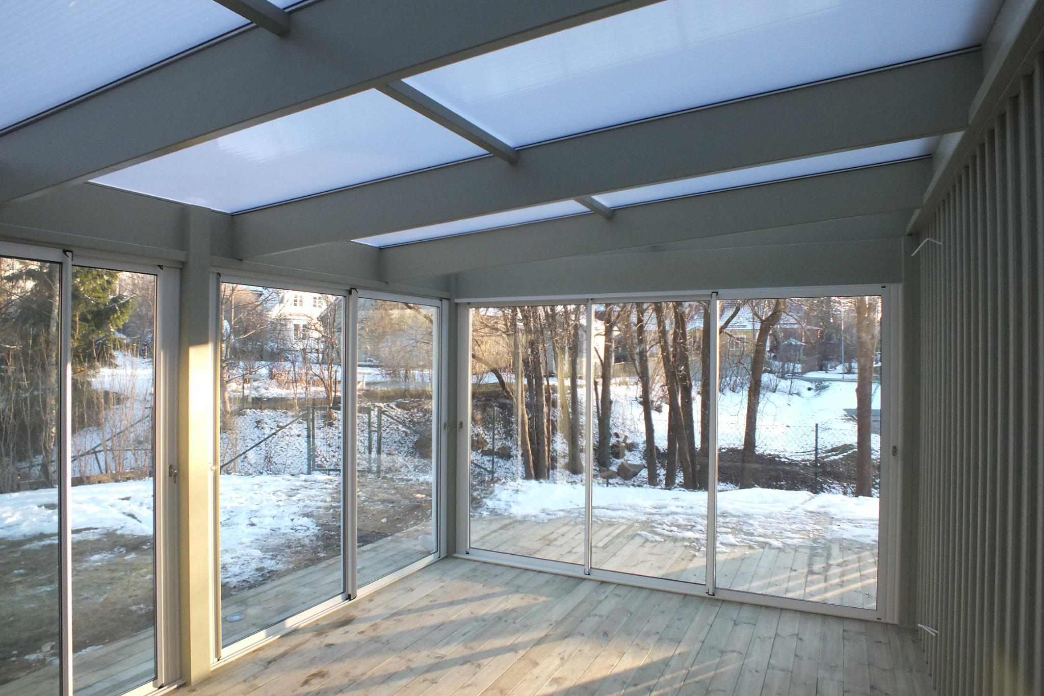 Nowoczesna zabudowa tarasu z drewnianą podłogą i przeszklonymi ścianami, widok na zimowy ogród. Przezroczysty dach z widoczną konstrukcją.