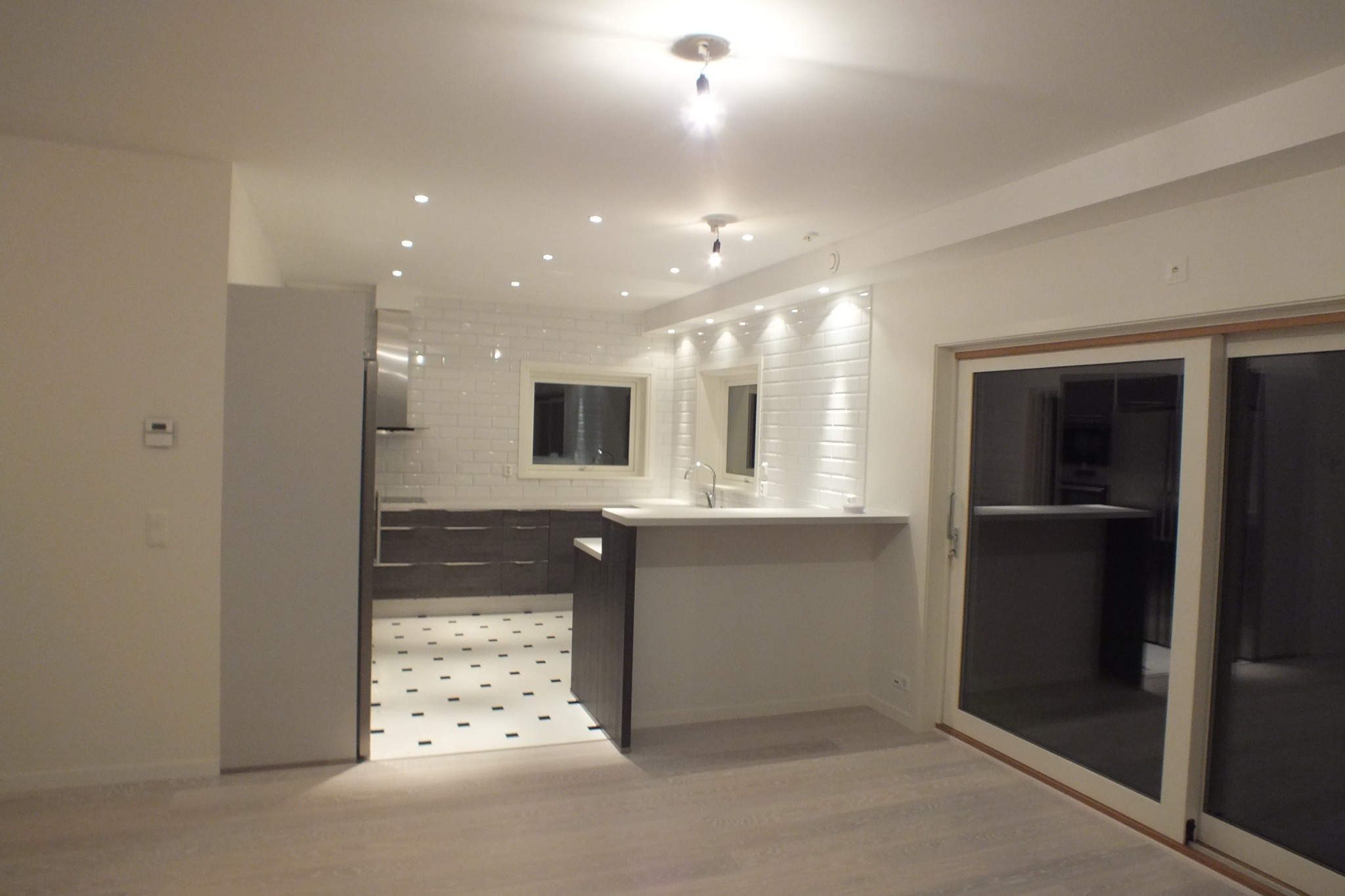 Jasne wnętrze kuchni z białymi płytkami na ścianach, ciemnymi szafkami i podłogą w biało-czarne wzory. Widoczne przesuwne drzwi i oświetlenie punktowe.