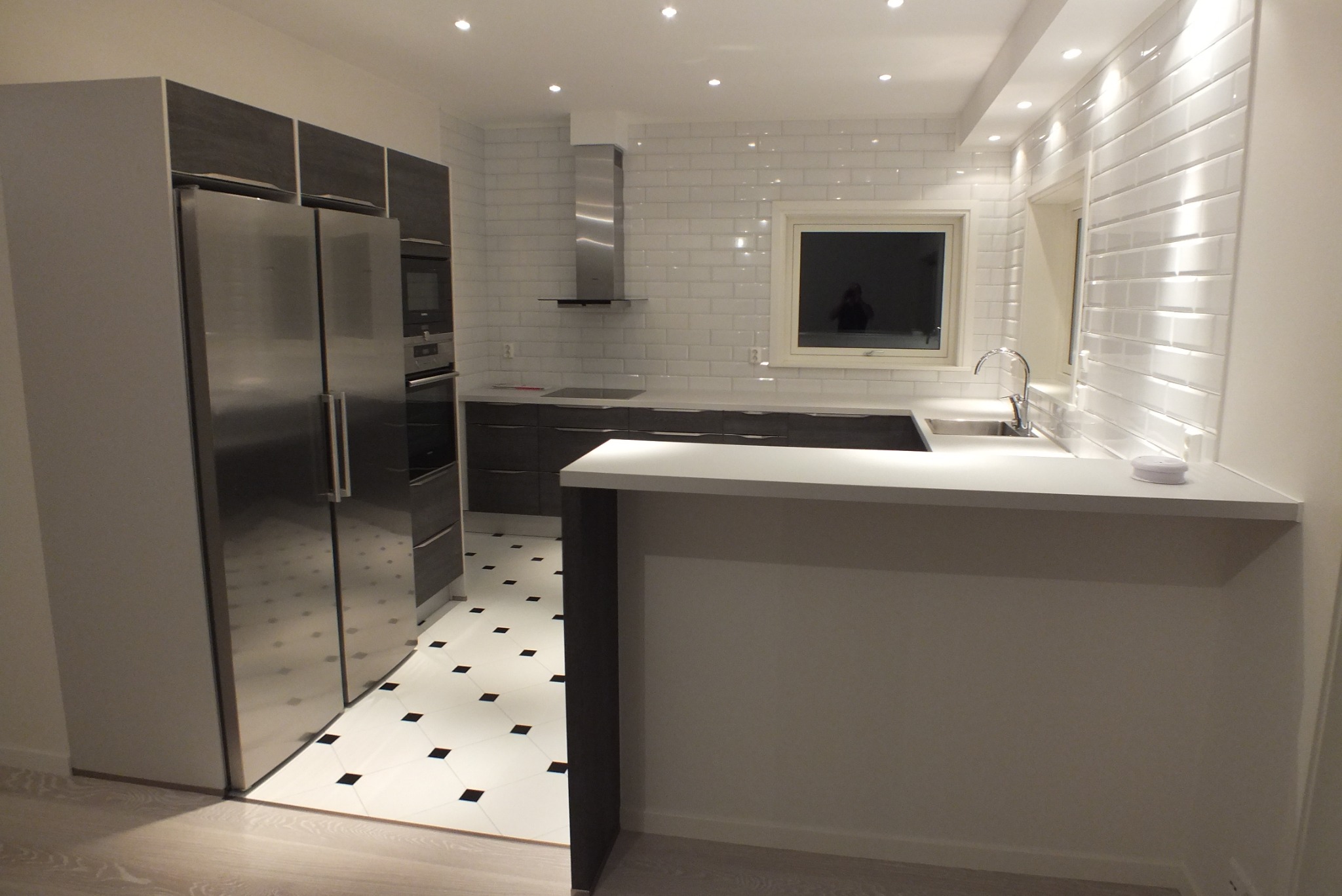 Nowoczesna, jasna kuchnia z białymi płytkami imitującymi cegłę, srebrną lodówką i czarno-białą podłogą w romby. Minimalistyczny design z oświetleniem punktowym.