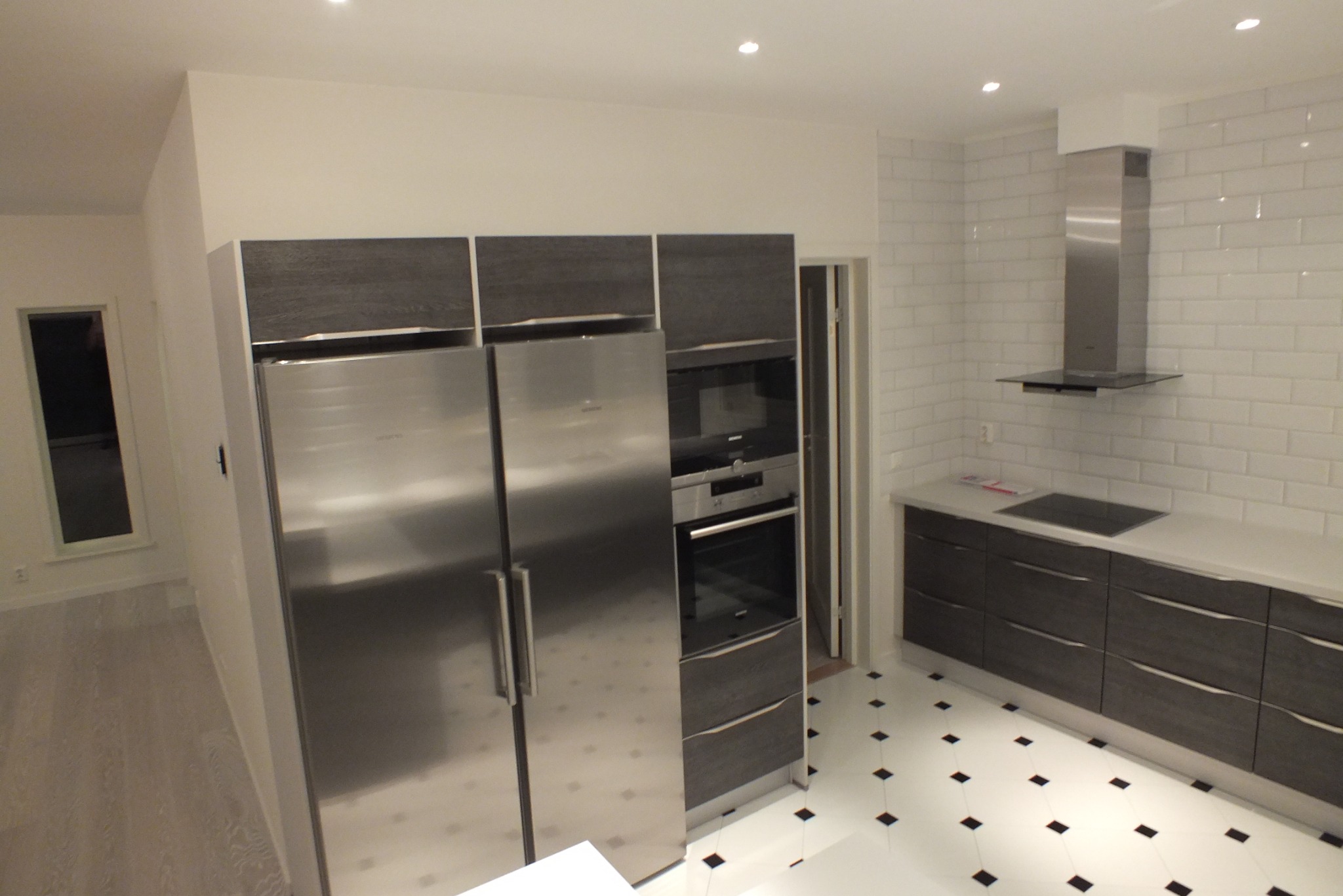 Nowoczesna, jasna kuchnia z białymi kaflami, zabudową w odcieniach szarości, stalową lodówką side-by-side i czarno-białą podłogą w karo.