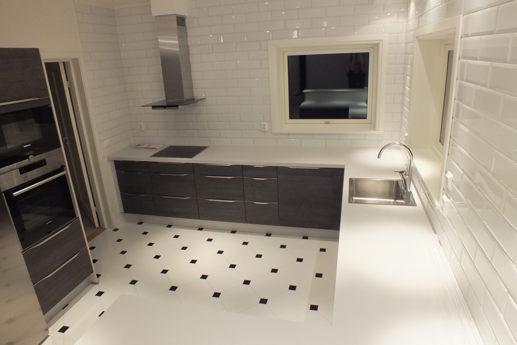 Nowoczesna kuchnia z białymi płytkami i ciemnymi szafkami. Widoczny zlew, okap, piekarnik i płyta indukcyjna. Podłoga biała w czarne kropki.