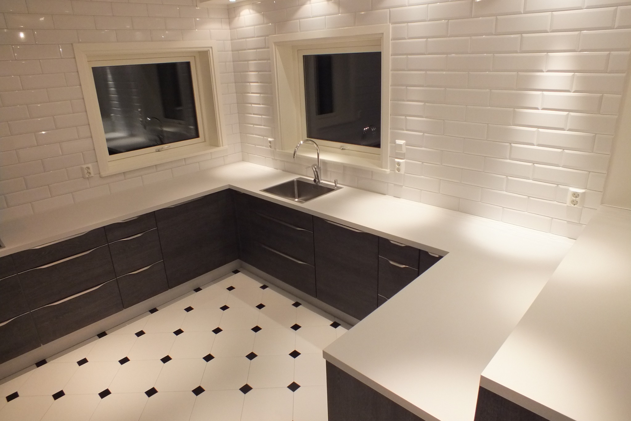 Nowoczesna kuchnia po remoncie: białe płytki na ścianach, ciemne szafki z delikatnymi uchwytami i biały blat. Na podłodze białe płytki z czarnymi akcentami.