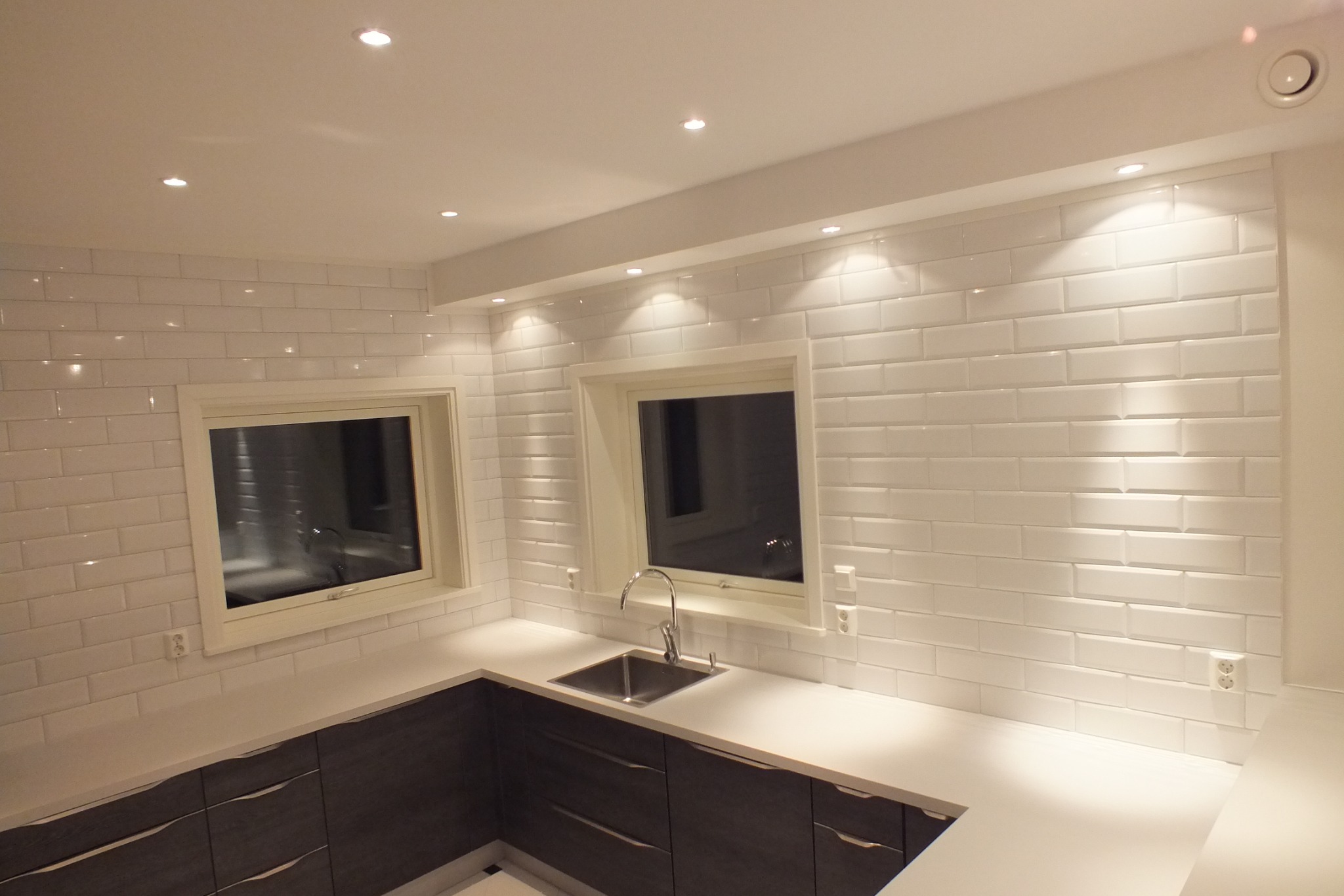 Nowoczesna kuchnia z białymi, cegiełkowymi płytkami, ciemnymi szafkami i jasnym blatem. Dwa okna w białych ramach. Oświetlenie sufitowe punktowe.