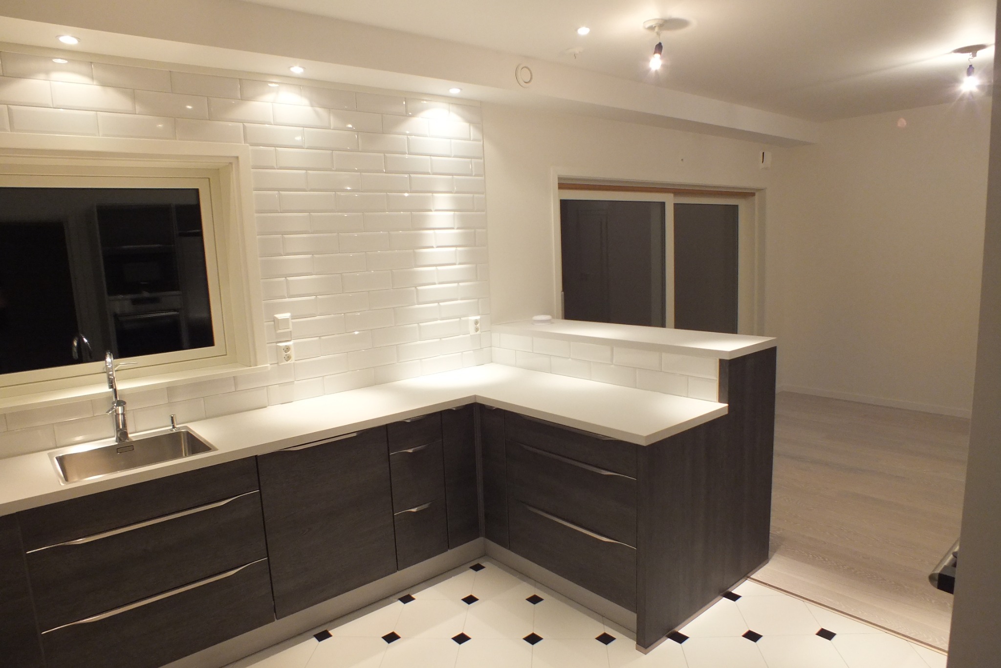 Nowoczesna kuchnia z ciemnymi szafkami i białym blatem, płytki metro na ścianie, okno z widokiem na zewnątrz, biało-czarna podłoga i oświetlenie punktowe.